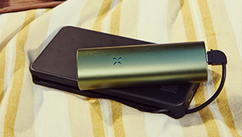 Un vaporizador Pax Flow verde elegante conectado a un cargador portátil negro mediante cable USB, descansando sobre una toalla a rayas amarillas y blancas para una carga conveniente en movimiento.