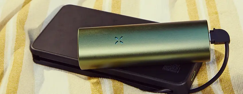 Un vaporizador Pax Flow verde elegante conectado a un cargador portátil negro mediante cable USB, descansando sobre una toalla a rayas amarillas y blancas para una carga conveniente en movimiento.