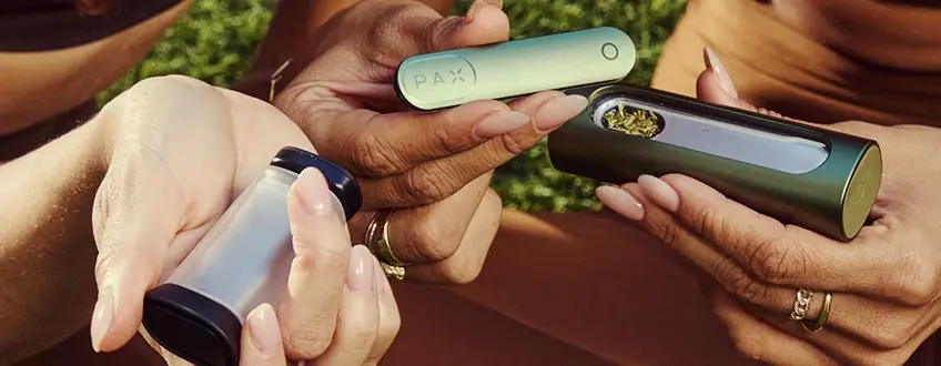 Dos manos colaborando para llenar el vaporizador Pax Flow con cannabis molido de un pequeño grinder, mostrando el proceso de carga fácil al aire libre sobre hierba verde.