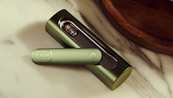 Primer plano del vaporizador Pax Flow abierto que revela su cámara interna llena de cannabis, sostenido por una mano en un entorno al aire libre casual con luz natural.