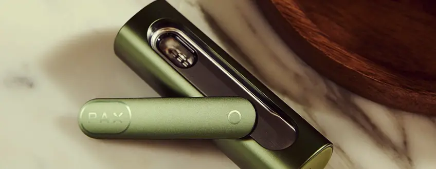 Primer plano del vaporizador Pax Flow abierto que revela su cámara interna llena de cannabis, sostenido por una mano en un entorno al aire libre casual con luz natural.