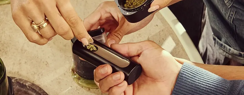 Una mano rellenando la cámara del vaporizador Pax Flow con cannabis molido fresco usando una pequeña cuchara de un grinder, colocado sobre una mesa de mármol para una recarga precisa.