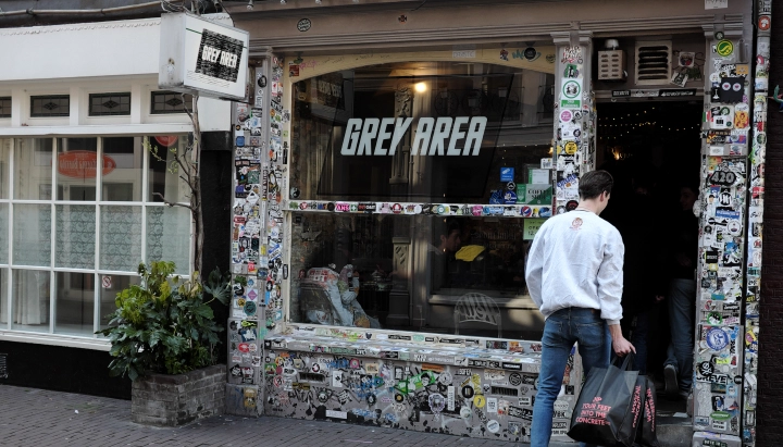 Entrada de un coffeeshop de cannabis en los Países Bajos con un cartel de Grey Area y una fachada cubierta de pegatinas.