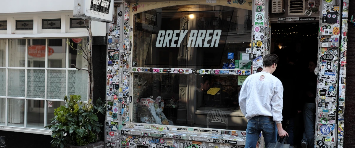 Entrada de un coffeeshop de cannabis en los Países Bajos con un cartel de Grey Area y una fachada cubierta de pegatinas.