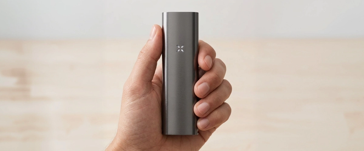 Vista de producto del vaporizador PAX Four en la mano Vaporizador PAX Four sostenido en una mano, mostrando su cuerpo fino y mate, su tamaño compacto y el indicador LED en forma de pétalos en la parte frontal