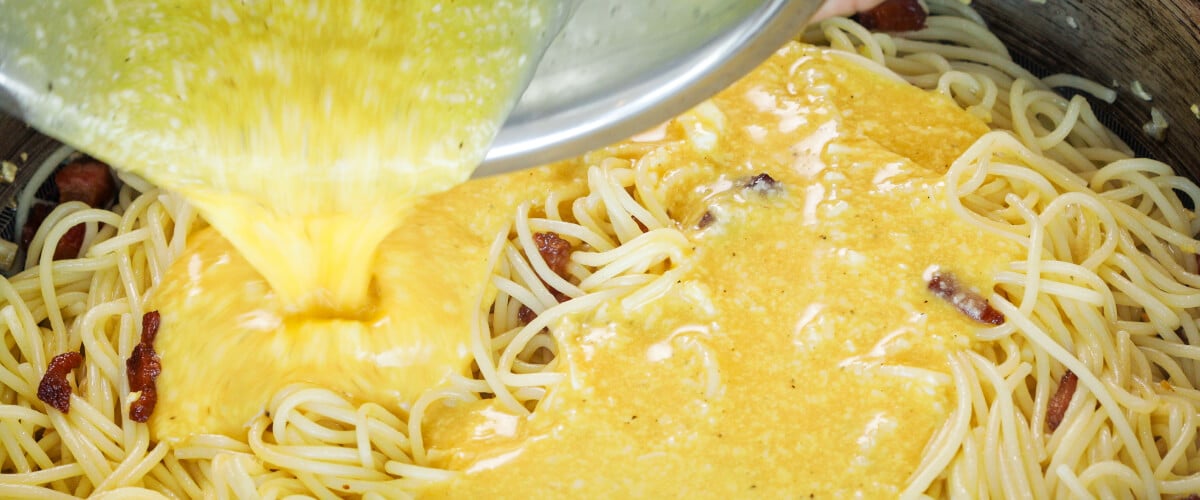 Vertiendo la mezcla de huevo y queso sobre espaguetis calientes en una sartén, quinto paso para una carbonara infusionada con cannabis
